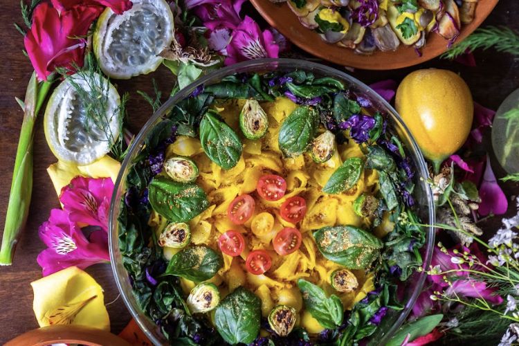 A glass bowl of greens, basil, cherry tomatoes and squash ribbons surrounded by flowers and fruit