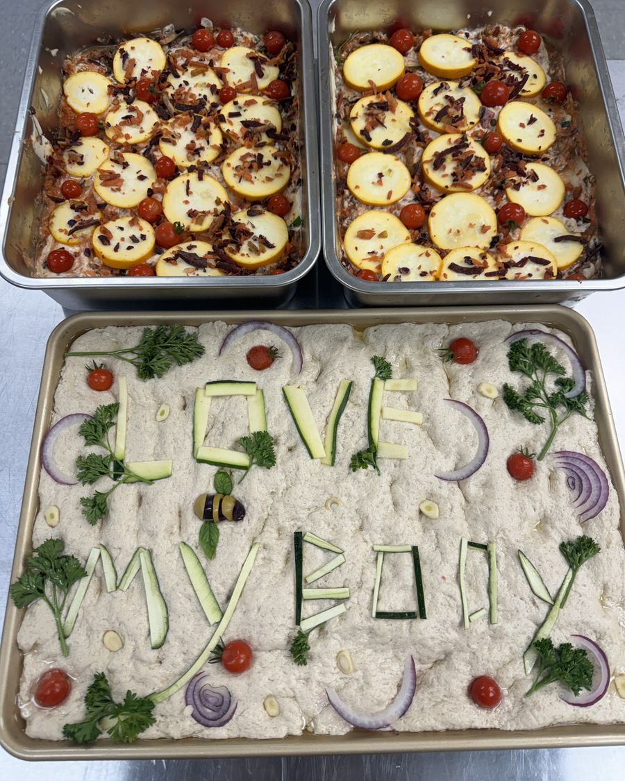 Focaccia decorated with vegetables spelling Love My Body, beside two trays of baked summer squash